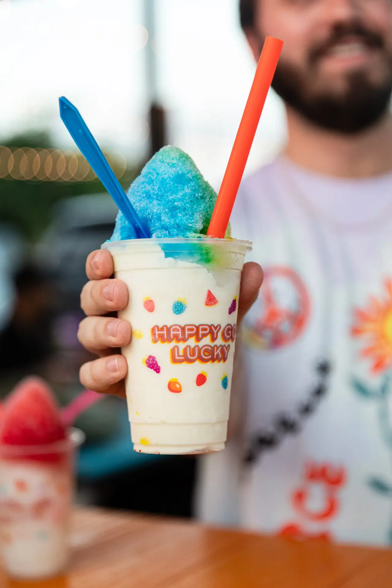 Hand holding a cup of rainbow shaved ice with blue and orange straws and colorful decorations on the cup surface.