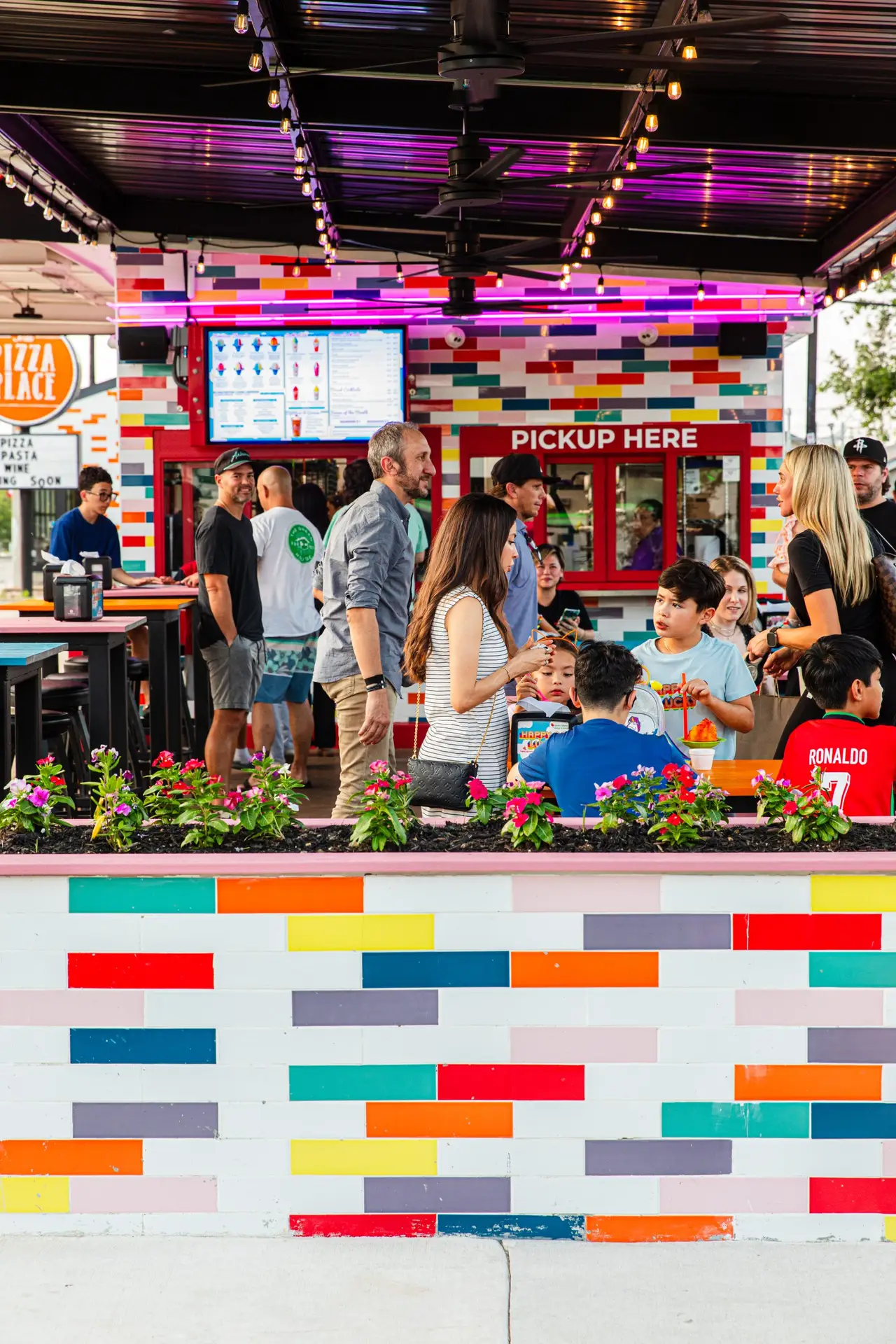 Outdoor colorful food stall with a striped rainbow wall, menu board, and people waiting for orders under string lights at dusk.